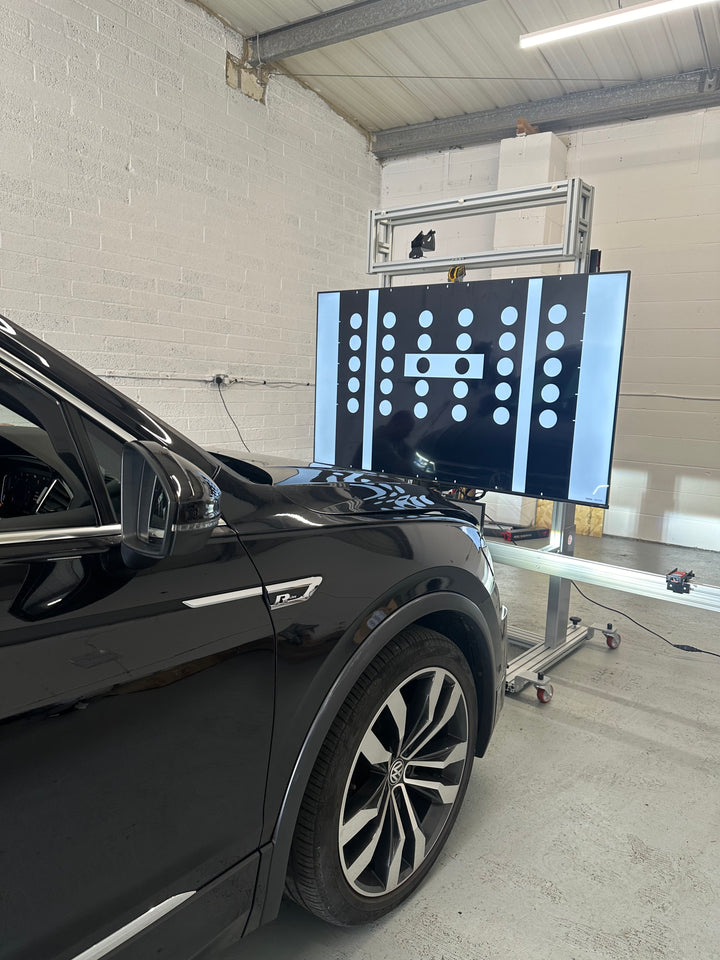 Black Volkswagen car parked in front of a tall stand holding a black board of white dots and vertical bars in a workshop
