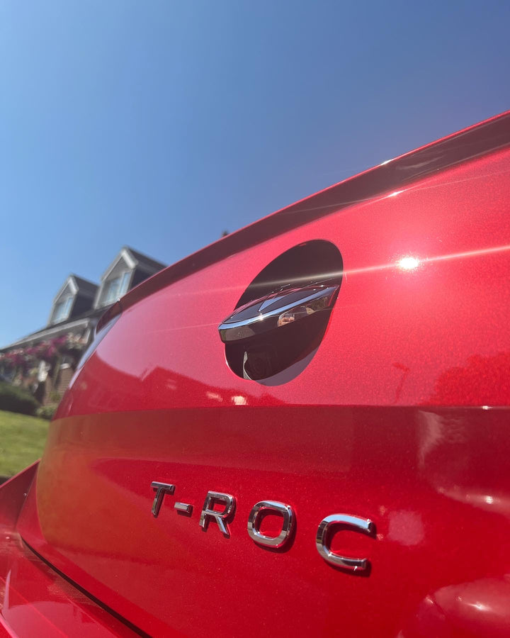 Red car tailgate with a “T-ROC” badge and a round emblem tilted open to show a small camera lens behind it