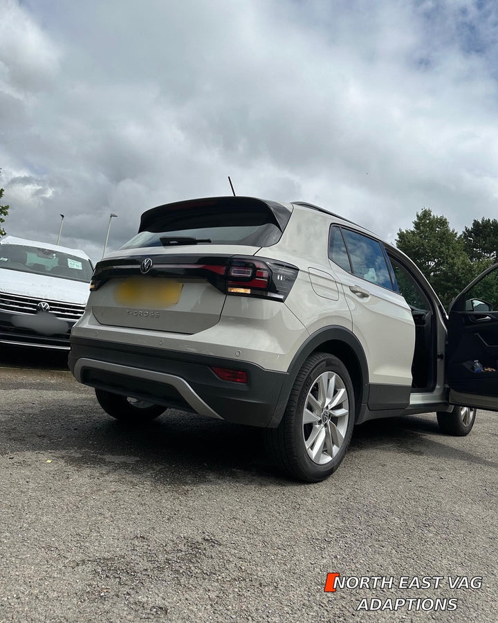 Rear of a cream VW SUV with a “T-CROSS” badge, parked in a car park with a door open; number plate blurred
