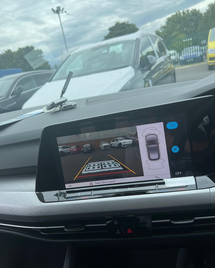 Dashboard touchscreen showing a rear camera view of a car park with a dotted floor board, guide lines and a top-down car icon